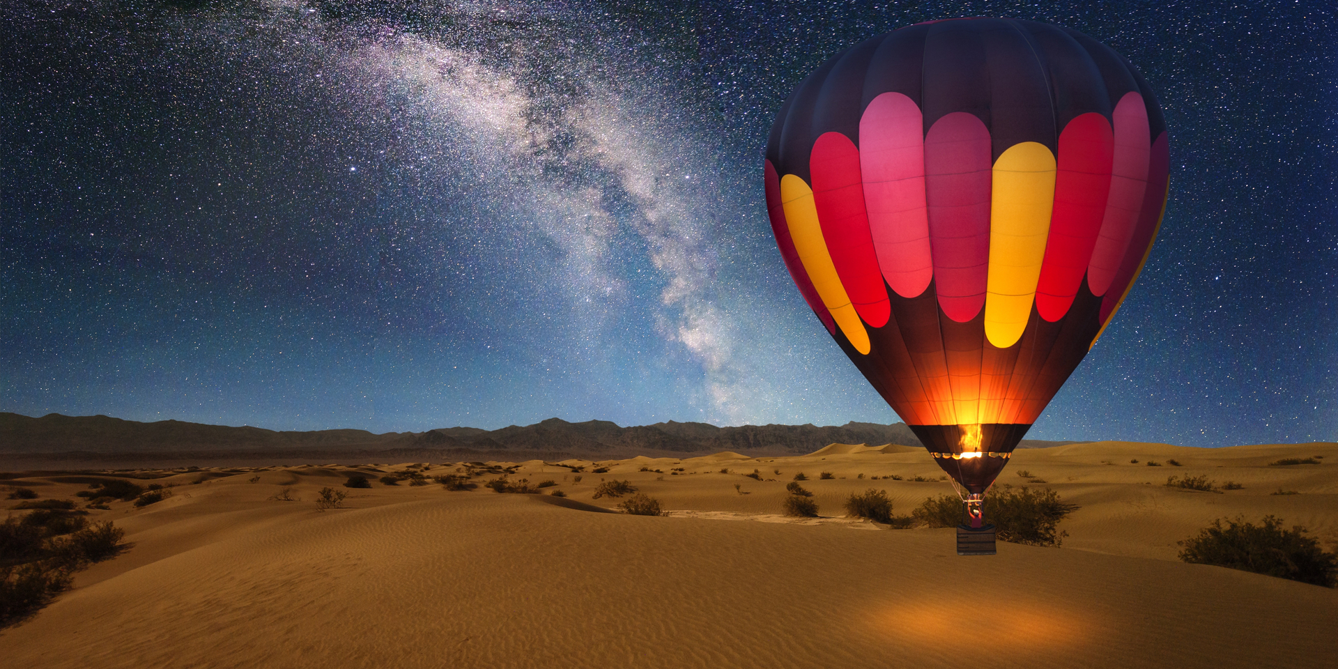 A majestic hot air balloon soars under the stars of the Milky Way, over the desert - Mesquite Dunes of Death Valley National Park. Moonlight provides luminosity showing the patterns and shapes of the desert landscape.