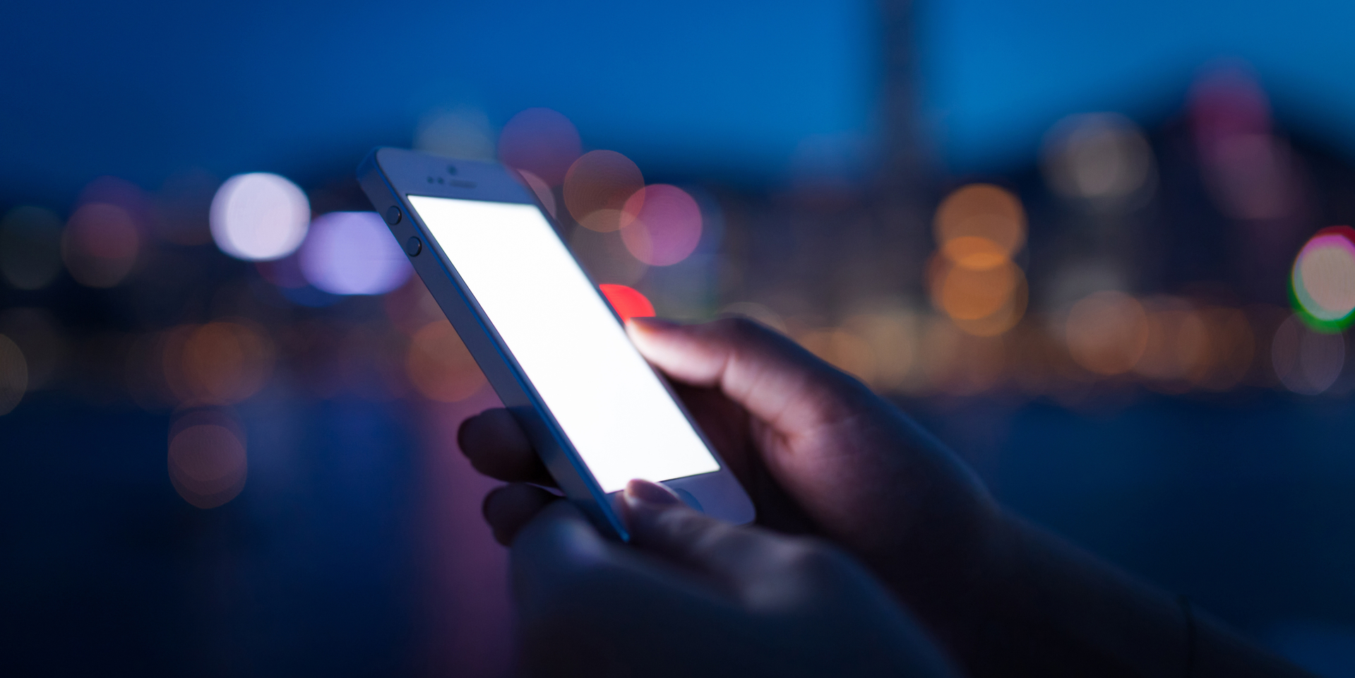 Human hands using smartphone in modern city, with blurry Hong Kong skyline at night time.