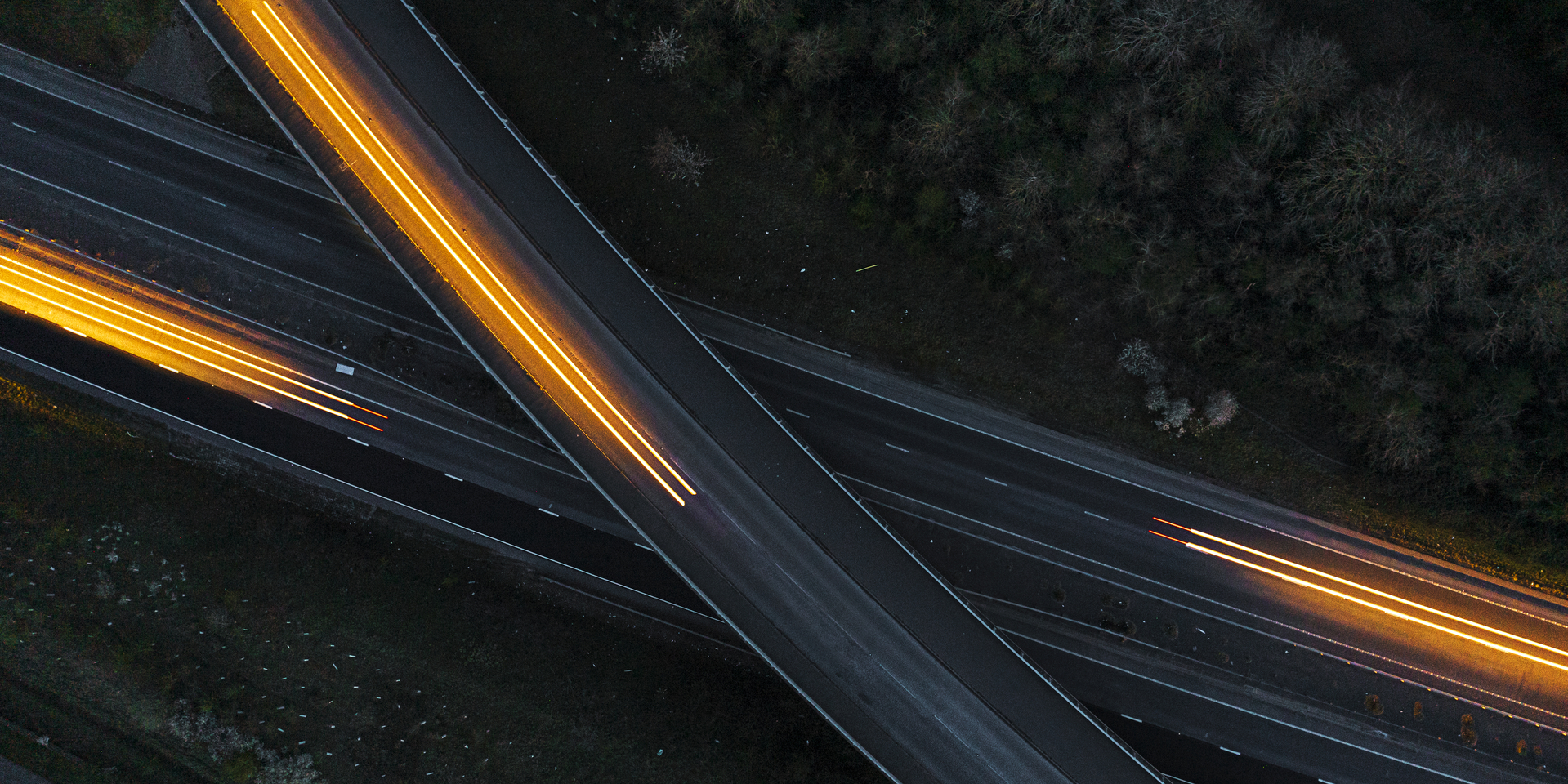 A long exposure from a drone shows the light trails of three cars moving in different directions on a road at night. One road passes over the main road
