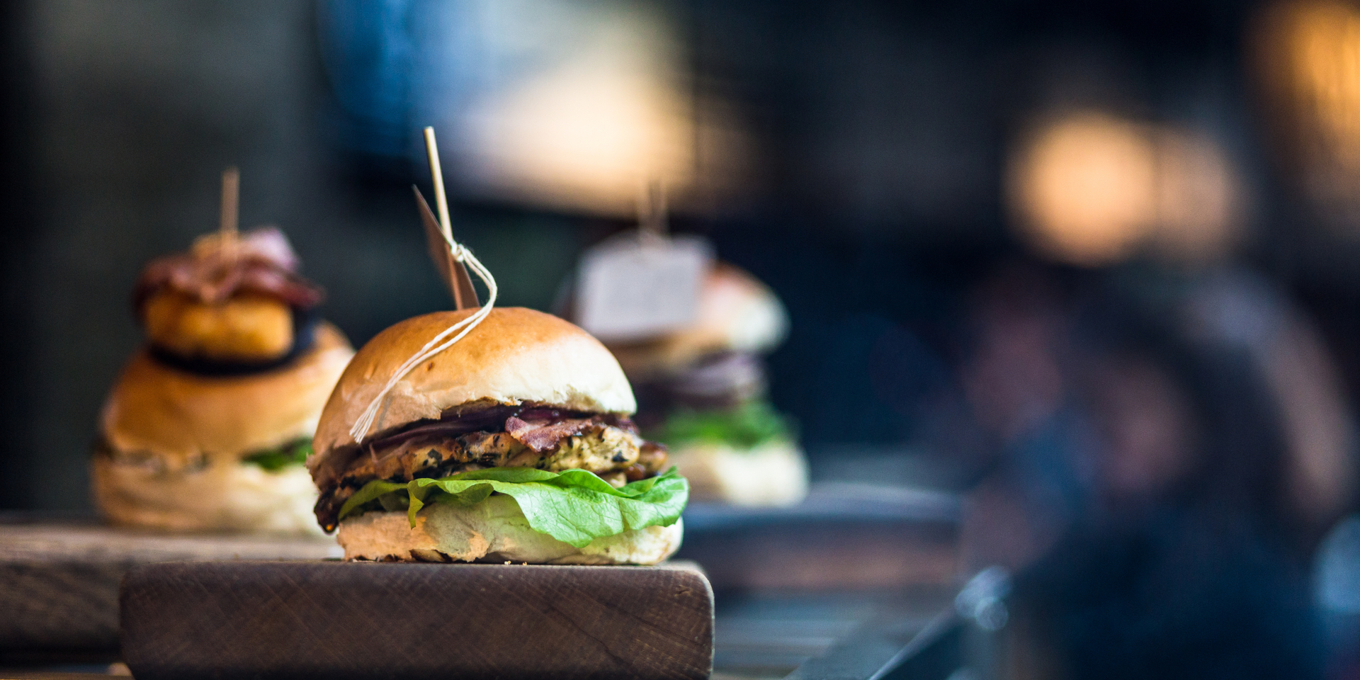 Close up image of a freshly flame grilled burger inside a bun, wilth lettuce, red onion, melted cheese and tomato. There are a few more burgers defocused in the background. The burger rests on a wooden board and the background is pleasantly defocused, leaving plenty of room for copy space. Horizontal colour image processed from an original RAW file, 