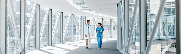Wide angle of two healthcare workers walking on covered glass bridge.